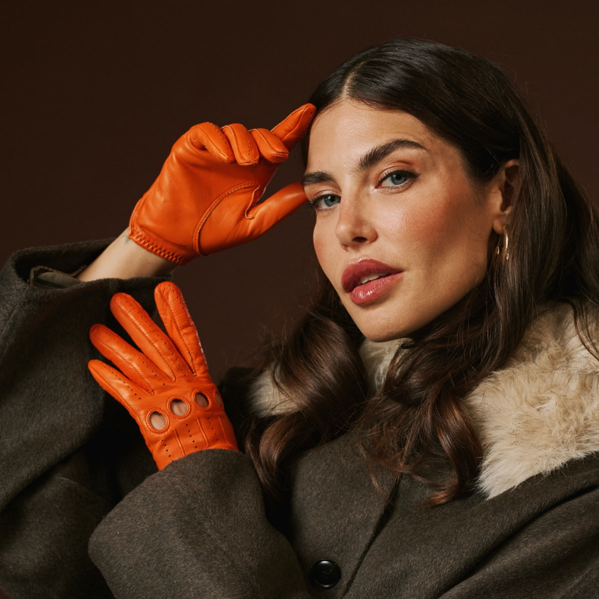 Woman wearing orange leather gloves and a brown coat against a dark background