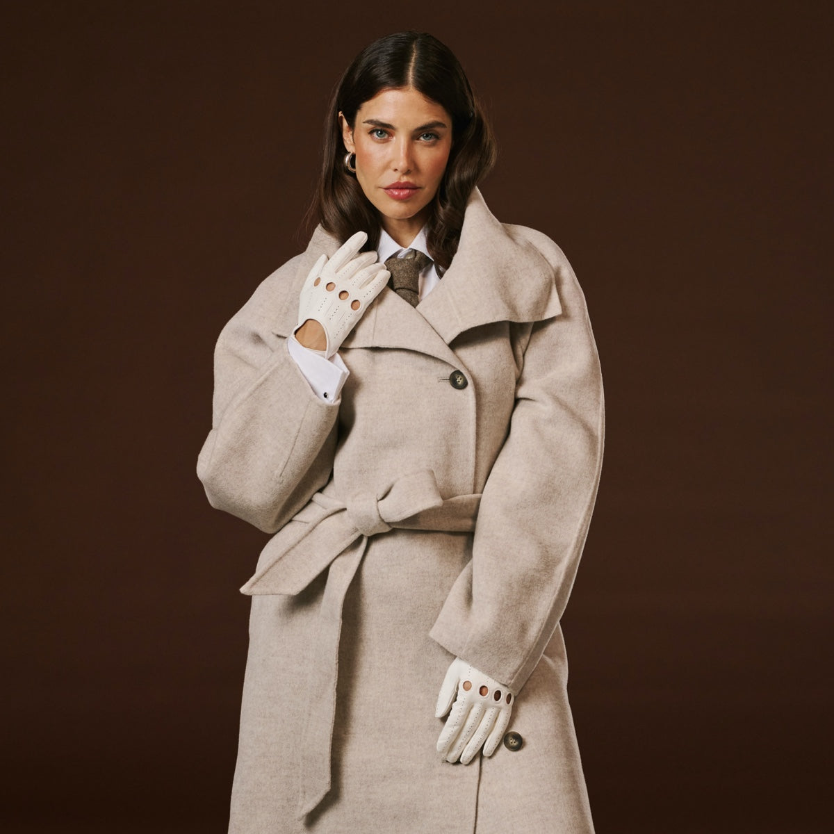 Woman wearing a beige trench coat and white leather  gloves against a brown background