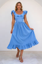 Woman in a light blue midi dress with oval neckline and smocked and ruffled off-the-shoulder sleeves against a white background.