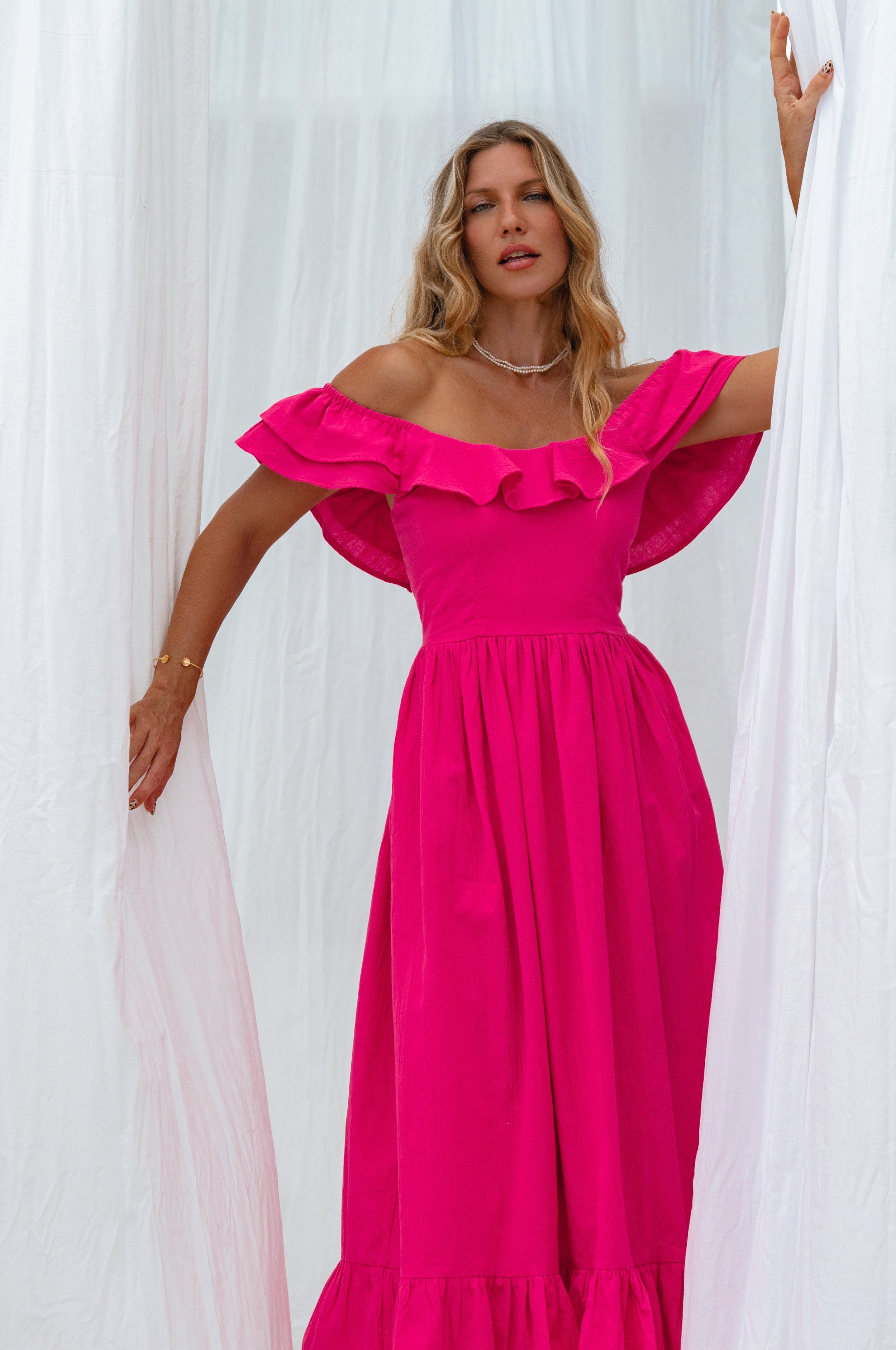 Woman in a bright pink midi dress with oval neckline and smocked and ruffled off-the-shoulder sleeves standing next to a white flower plant against a white curtain, front view