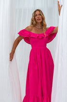 Woman in a bright pink midi dress with oval neckline and smocked and ruffled off-the-shoulder sleeves standing next to a white flower plant against a white curtain, front view