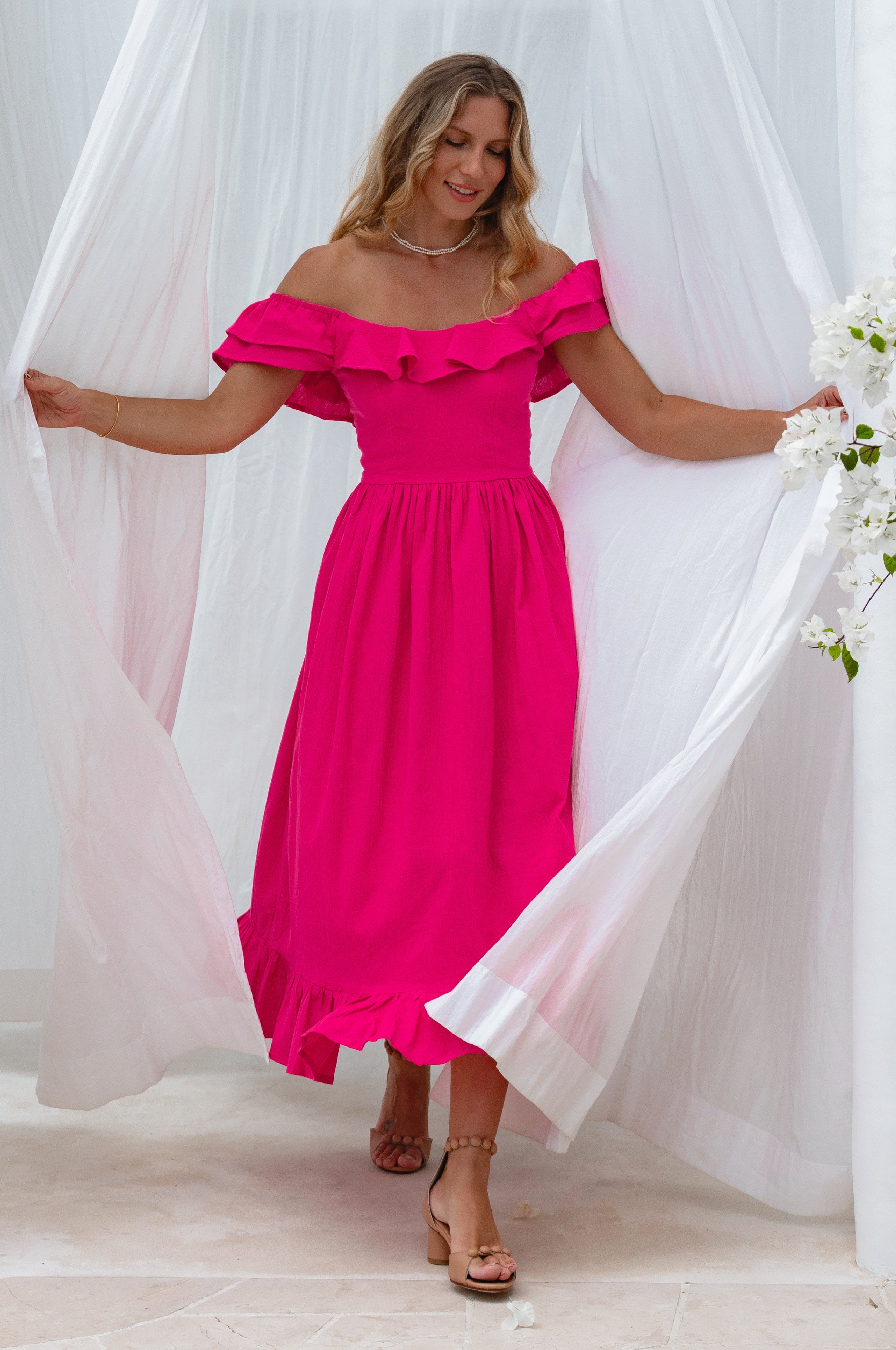 Woman in a bright pink midi dress with oval neckline and smocked and ruffled off-the-shoulder sleeves standing next to a white flower plant against a white curtain.