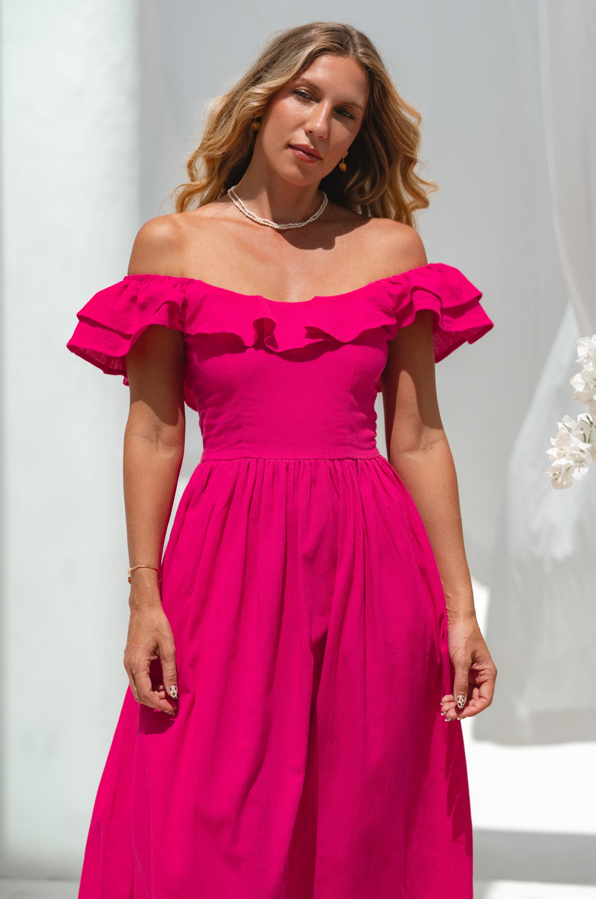 Woman in a bright pink midi dress with oval neckline and smocked and ruffled off-the-shoulder sleeves standing next to a white flower plant against a white curtain.