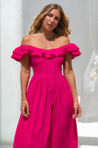 Woman in a bright pink midi dress with oval neckline and smocked and ruffled off-the-shoulder sleeves standing next to a white flower plant against a white curtain.