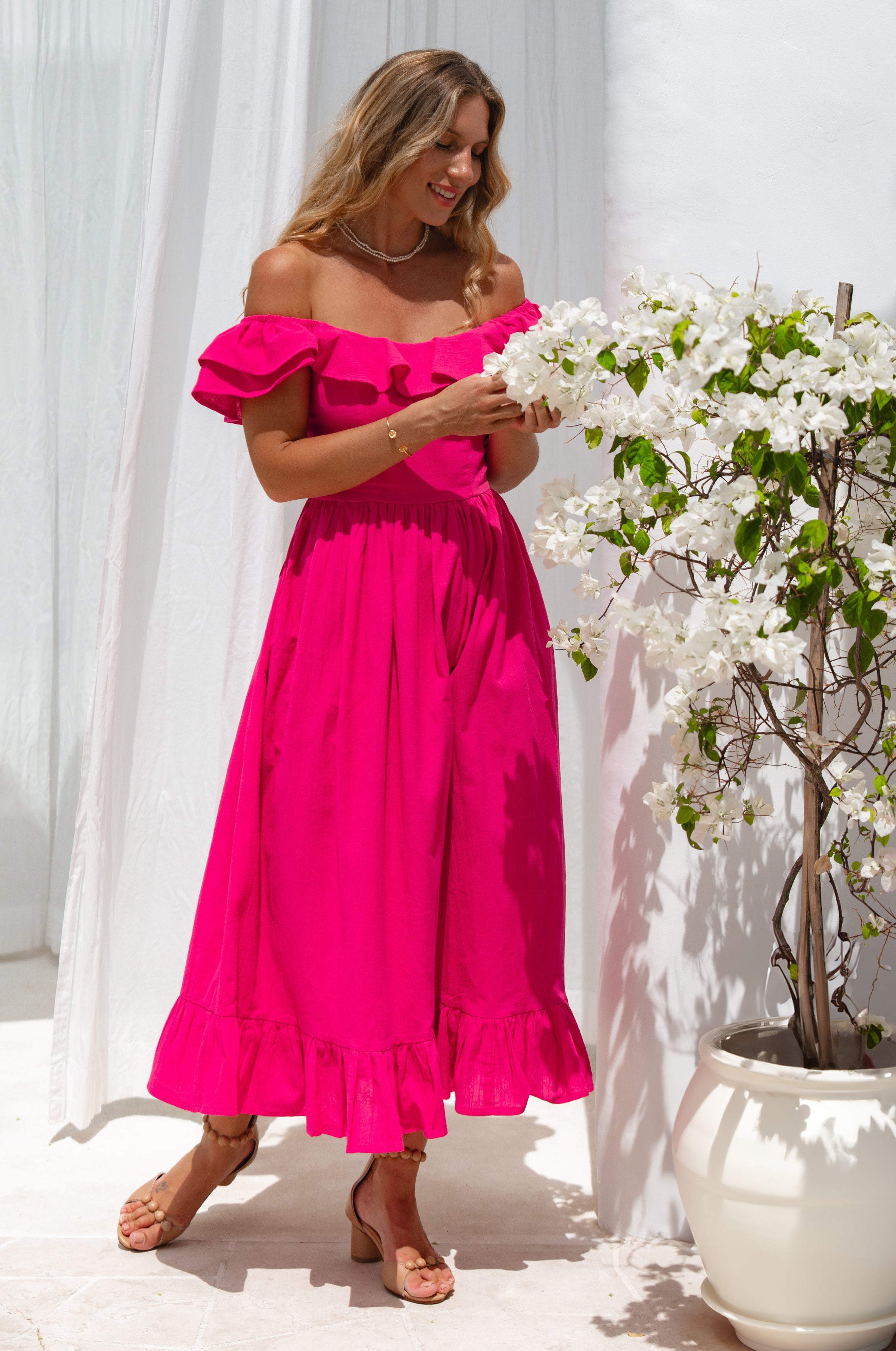 Woman in a bright pink midi dress with oval neckline and smocked and ruffled off-the-shoulder sleeves standing next to a white flower plant against a white curtain.