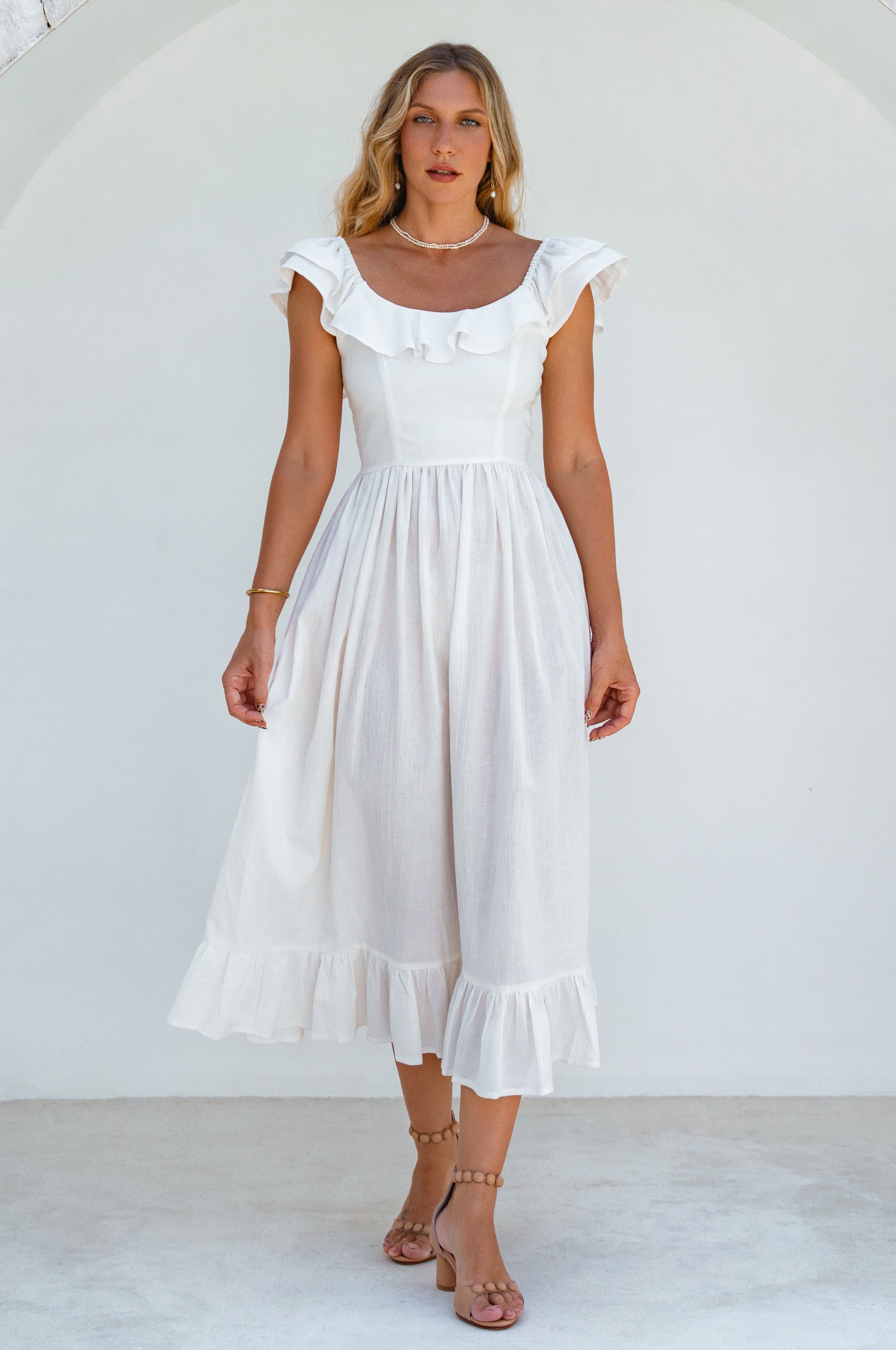 Woman in a white midi dress with oval neckline and smocked and ruffled off-the-shoulder sleeves against a white background, front view