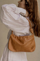 A woman wearing a white puff-sleeve blouse carries a tan leather shoulder bag with a knotted strap detail against a neutral beige background.
