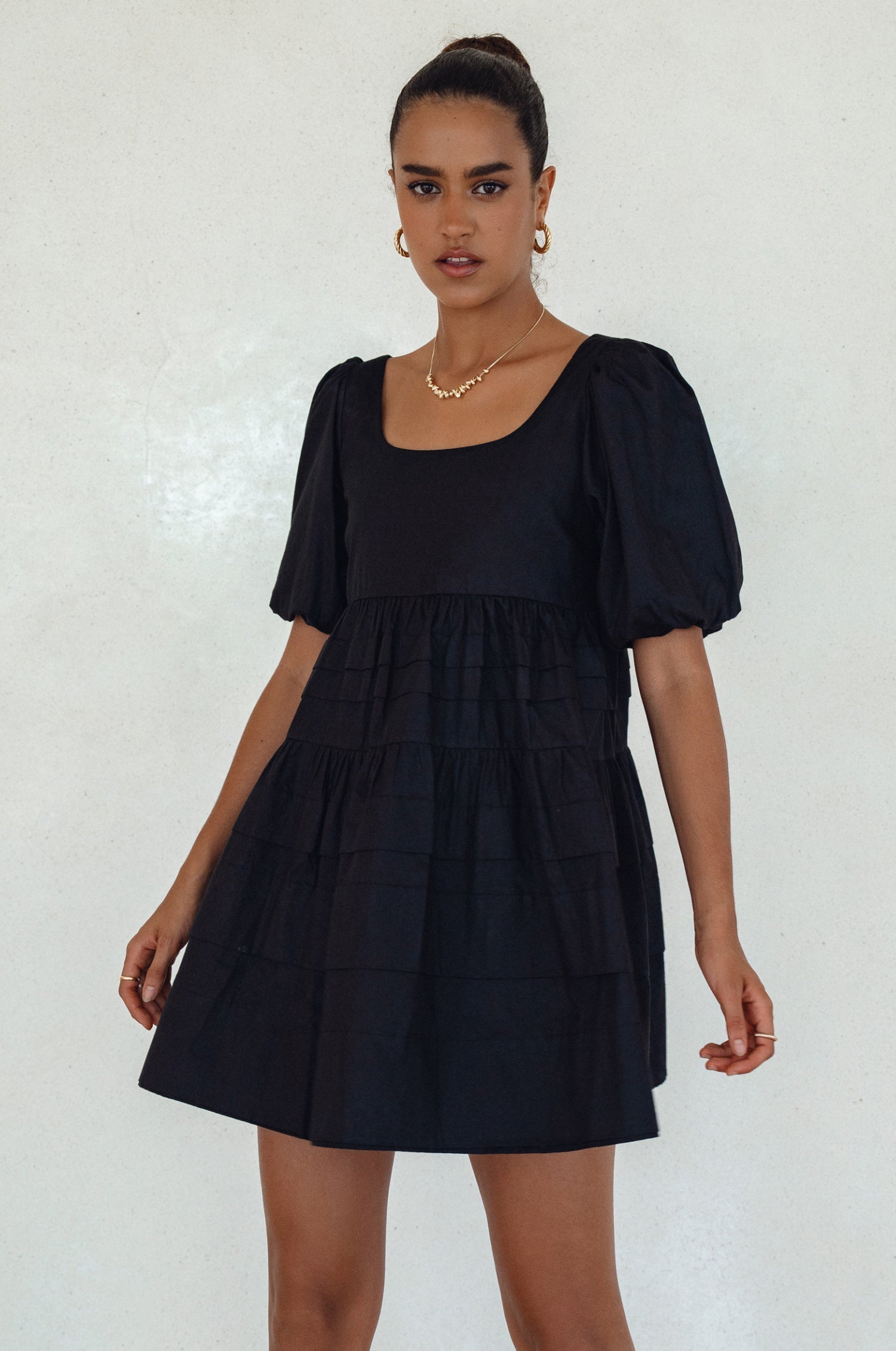 Woman wearing a black mini dress slightly voluminous elasticated sleeves, tiered folds on the hem and round neckline against a white background, front view