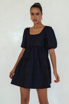 Woman wearing a black mini dress slightly voluminous elasticated sleeves, tiered folds on the hem and round neckline against a white background, front view