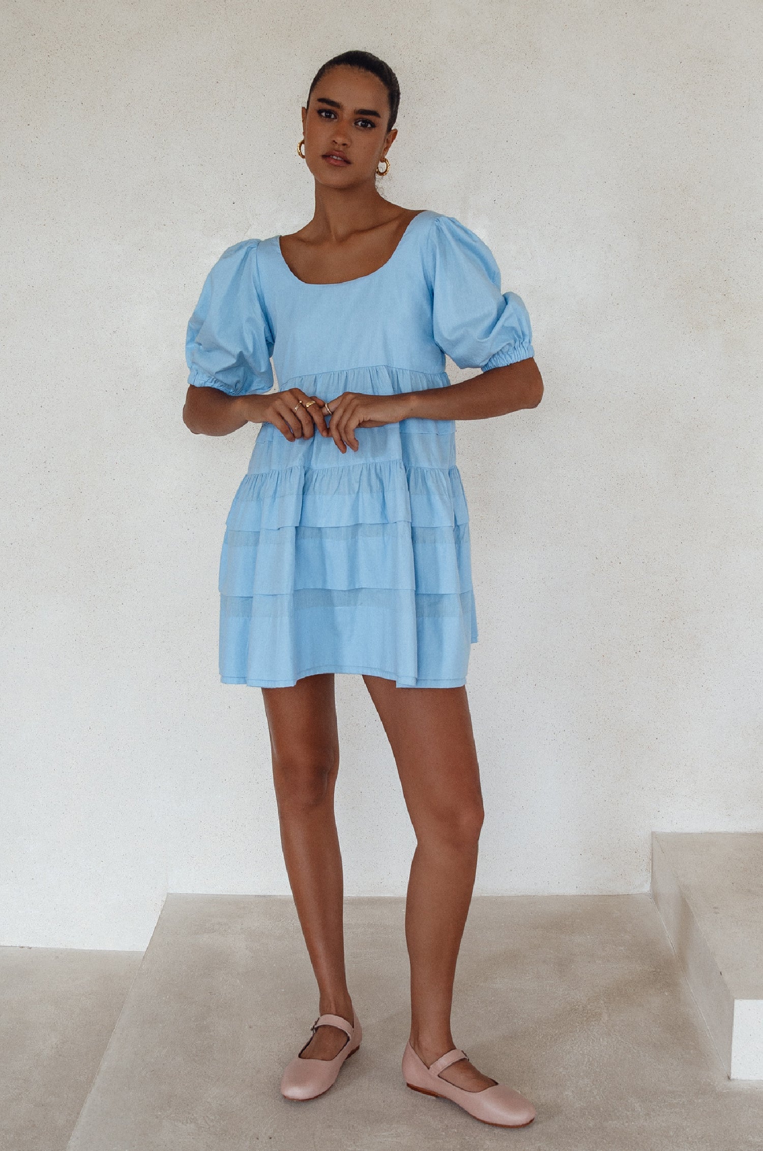 Woman wearing a light blue mini dress slightly voluminous elasticated sleeves, folds on the hem and round neckline.  against a white background, front view
