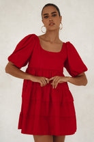 Woman wearing a red mini dress slightly voluminous elasticated sleeves, tiered folds on the hem and round neckline against a white background, front view