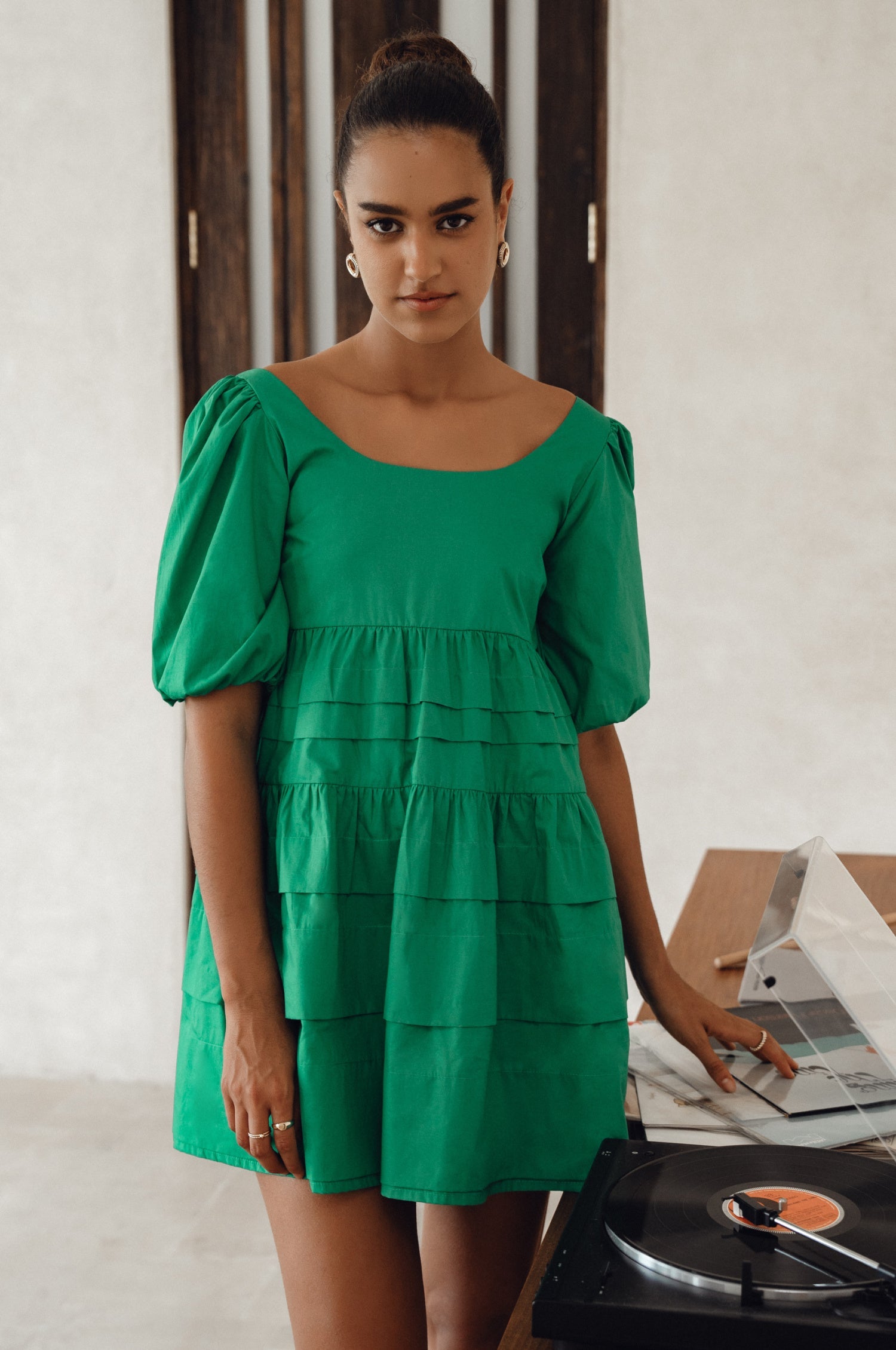 Woman wearing a green mini dress slightly voluminous elasticated sleeves, tiered folds on the hem and round neckline, front view