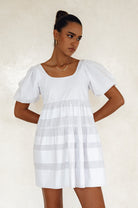 Woman wearing a white mini dress slightly voluminous elasticated sleeves, tiered folds on the hem and round neckline against a white background, front view