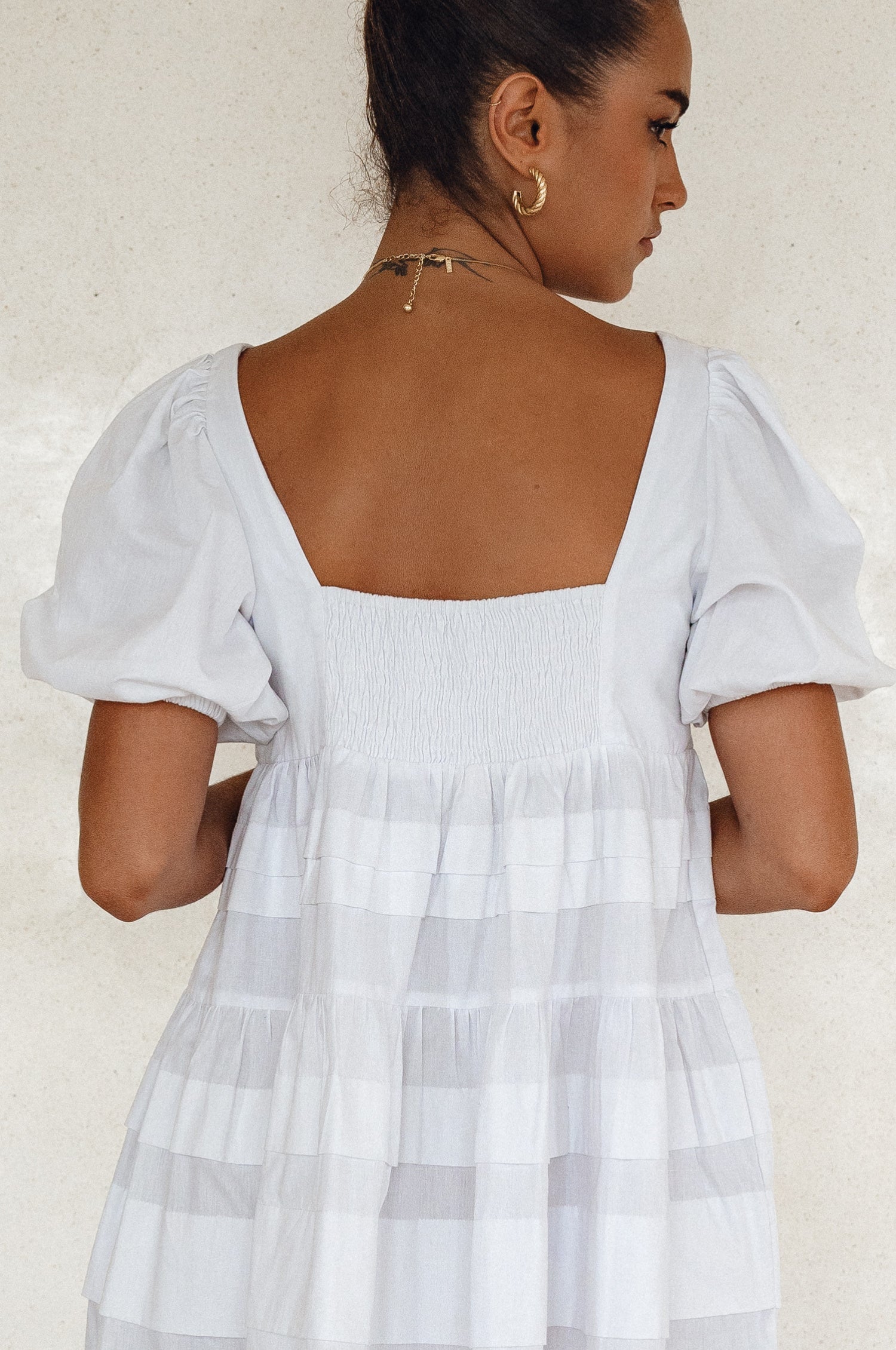 Woman wearing a white mini dress slightly voluminous elasticated sleeves, tiered folds on the hem and round neckline against a white background, back view