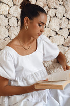 Woman wearing a white mini dress slightly voluminous elasticated sleeves, tiered folds on the hem and round neckline against reading a book against a stone background, front view