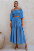 Woman wearing blue crinkle linen high-waist two-tier maxi skirt and matching cropped off-shoulder blouse against neutral background, front view. 