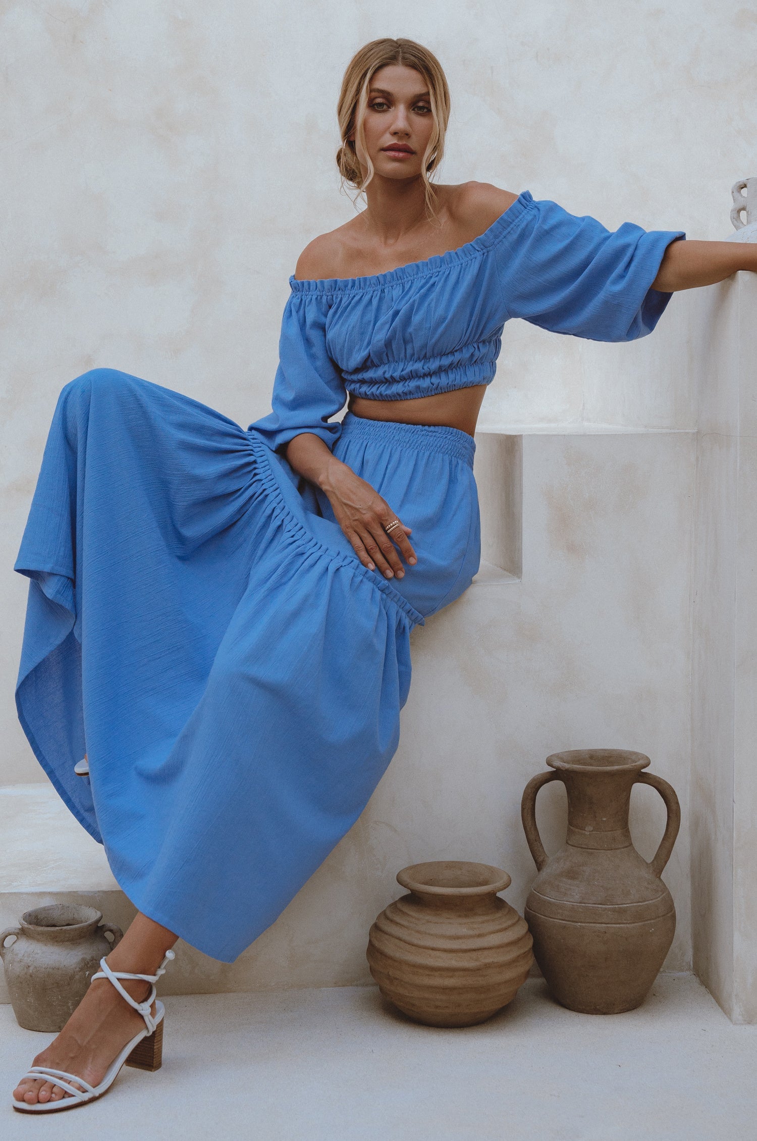 Woman wearing blue crinkle linen high-waist two-tier maxi skirt and matching cropped off-shoulder blouse against neutral background, front view. 