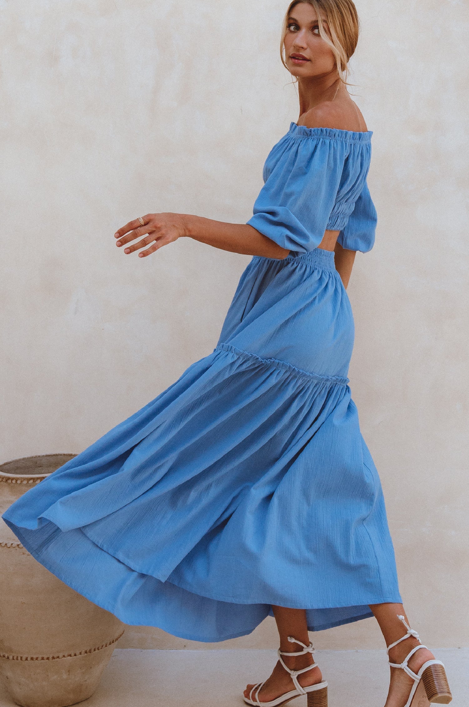 Woman wearing blue crinkle linen high-waist two-tier maxi skirt and matching cropped off-shoulder blouse against neutral background, side view. 