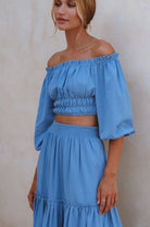 Woman wearing white crinkle linen high-waist two-tier maxi skirt and matching cropped off-shoulder blouse against neutral background, 3/4 view. 