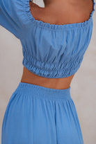 Woman wearing blue crinkle linen high-waist two-tier maxi skirt and matching cropped off-shoulder blouse against neutral background, back view. 
