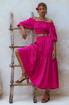 Woman wearing magenta crinkle linen high-waist two-tier maxi skirt and matching cropped off-shoulder blouse against neutral background, front view. 