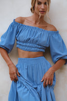 Woman wearing a blue linen cropped top with elasticated detailing at the waist and neckline worn off-the-shoulder against a neutral background, front view