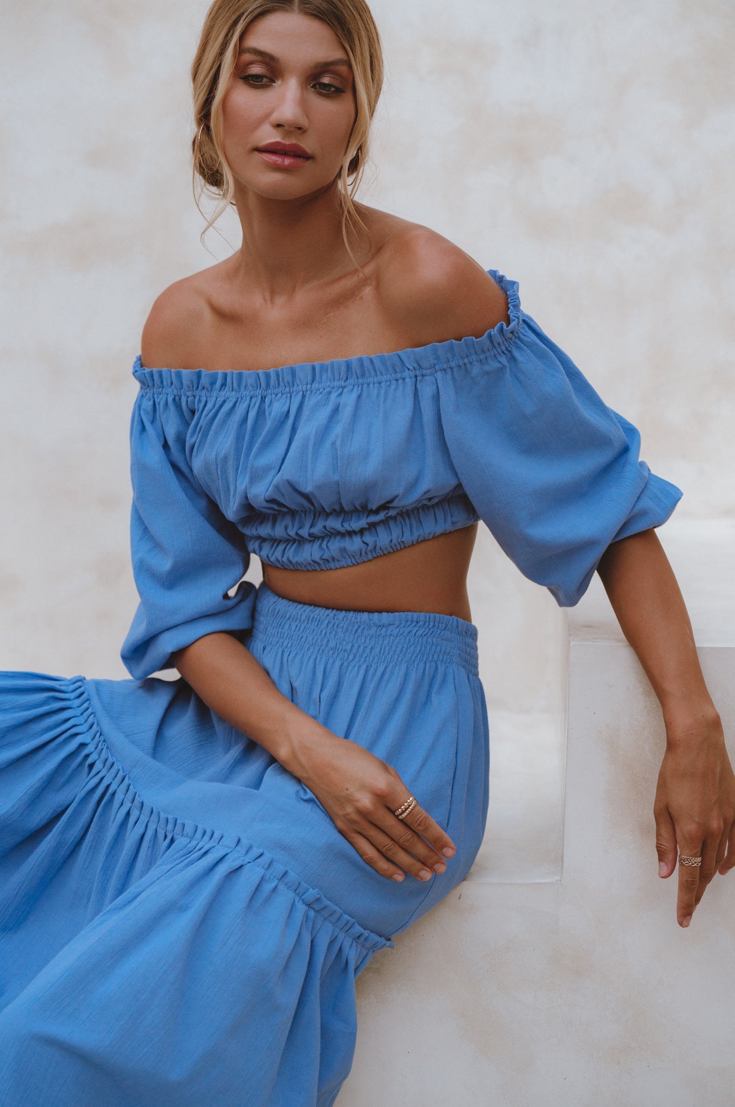 Woman wearing a blue linen cropped top with elasticated detailing at the waist and neckline worn off-the-shoulder and matching maxi skirt against a white background, 3/4 view