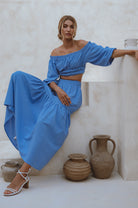 Woman wearing a blue linen cropped top with elasticated detailing at the waist and neckline worn off-the-shoulder and matching maxi skirt, front view