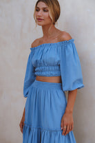 Woman wearing a blue linen cropped top with elasticated detailing at the waist and neckline worn off-the-shoulder and matching maxi skirt against a white background, 3/4 view