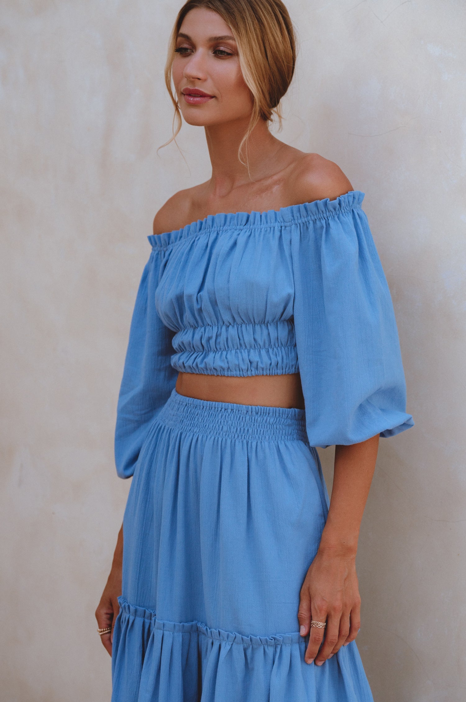 Woman wearing a blue linen cropped top with elasticated detailing at the waist and neckline worn off-the-shoulder and matching maxi skirt against a white background, 3/4 view
