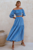 Woman wearing a blue linen cropped top with elasticated detailing at the waist and neckline worn off-the-shoulder and matching maxi skirt against a white background, front view