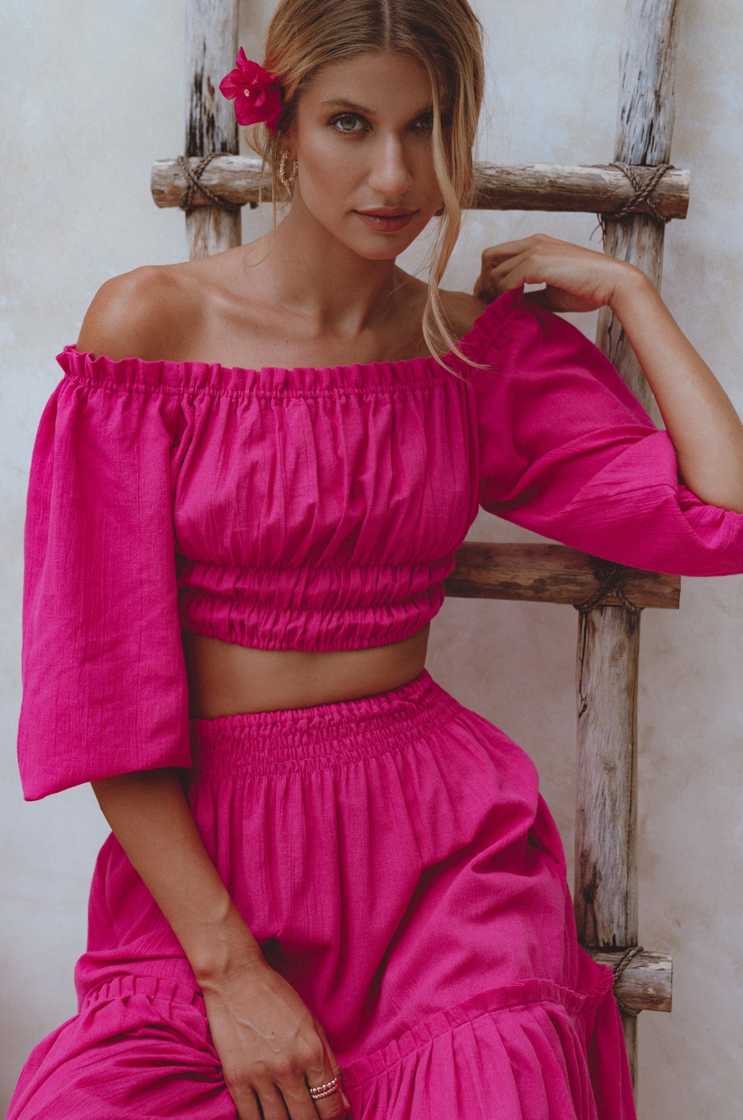Woman wearing a magenta linen cropped top with elasticated detailing at the waist and neckline worn off-the-shoulder against a neutral background, front view