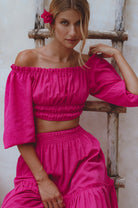 Woman wearing a magenta linen cropped top with elasticated detailing at the waist and neckline worn off-the-shoulder and matching maxi skirt against a white background, front view