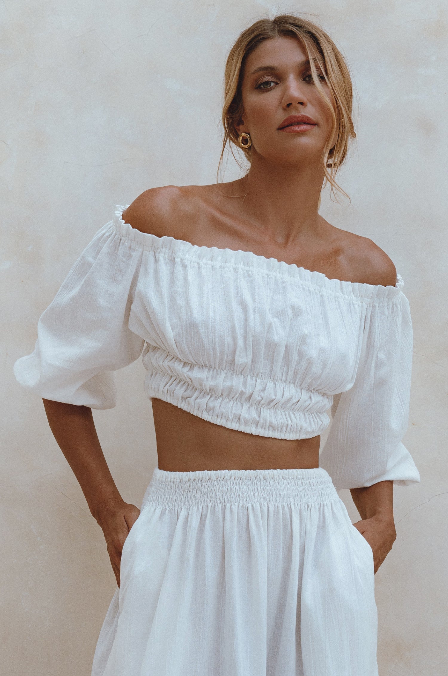 Woman wearing a white linen cropped top with elasticated detailing at the waist and neckline worn off-the-shoulder against a white background, front view