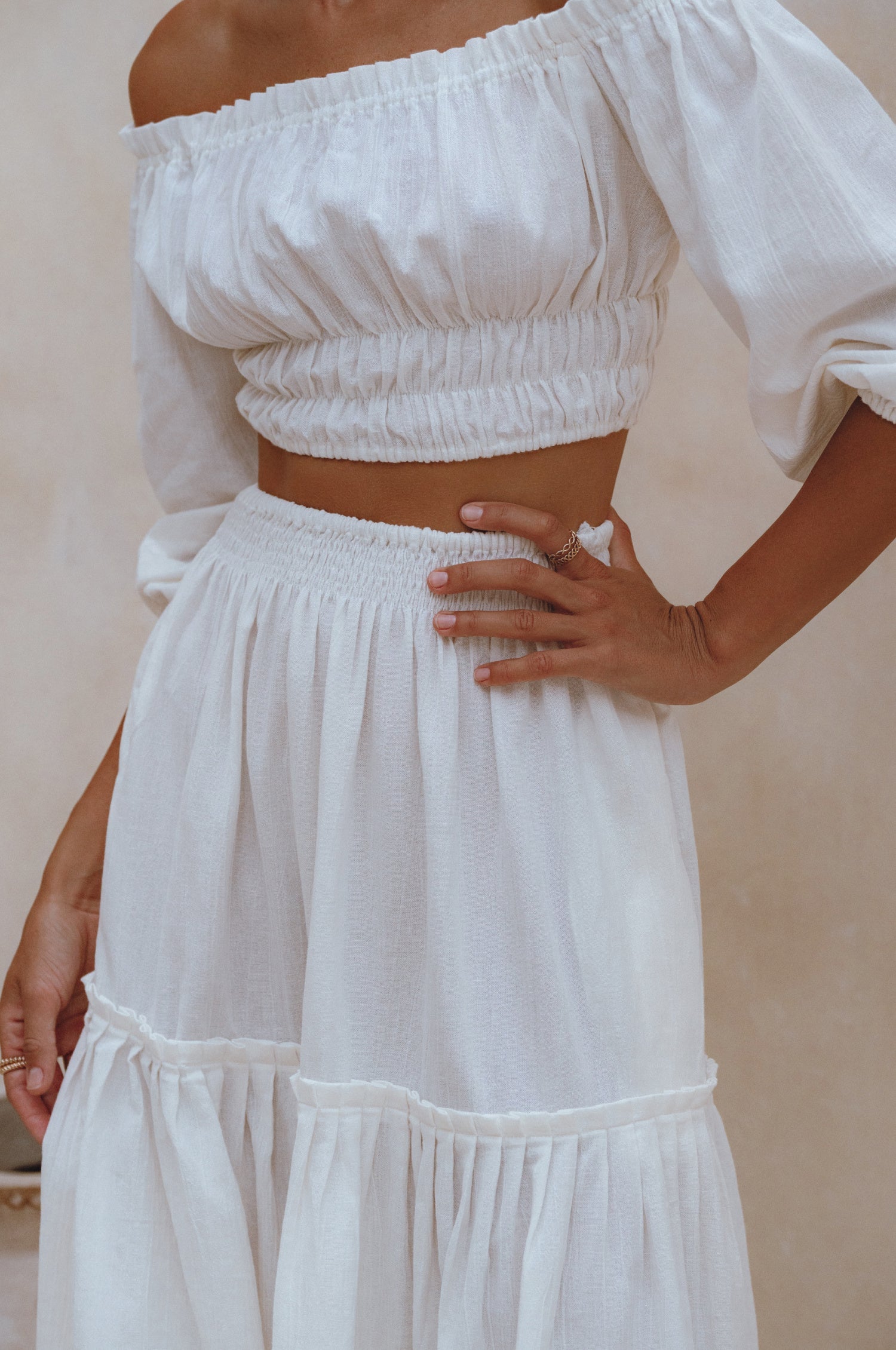 Woman wearing a white linen cropped top with elasticated detailing at the waist and neckline worn off-the-shoulder against a white background, 3/4 view
