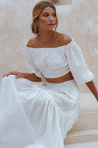 Woman wearing a white linen cropped top with elasticated detailing at the waist and neckline worn off-the-shoulder against a neutral background, front view