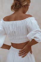 Woman wearing a white linen cropped top with elasticated detailing at the waist and neckline worn off-the-shoulder against a white background, back view