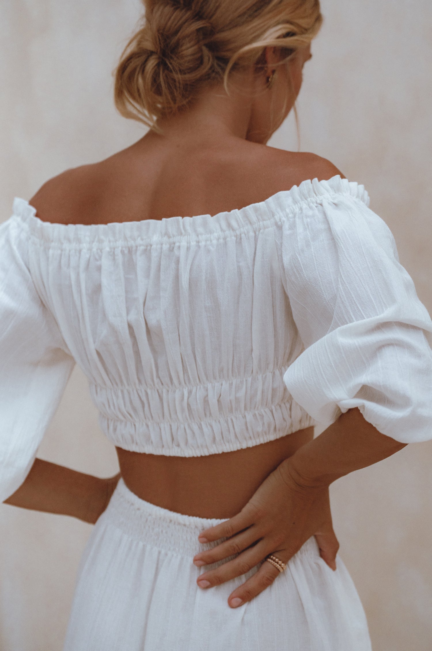 Woman wearing a white linen cropped top with elasticated detailing at the waist and neckline worn off-the-shoulder against a white background, back view