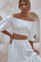 Woman wearing a white linen cropped top with elasticated detailing at the waist and neckline worn off-the-shoulder against a white background, front view