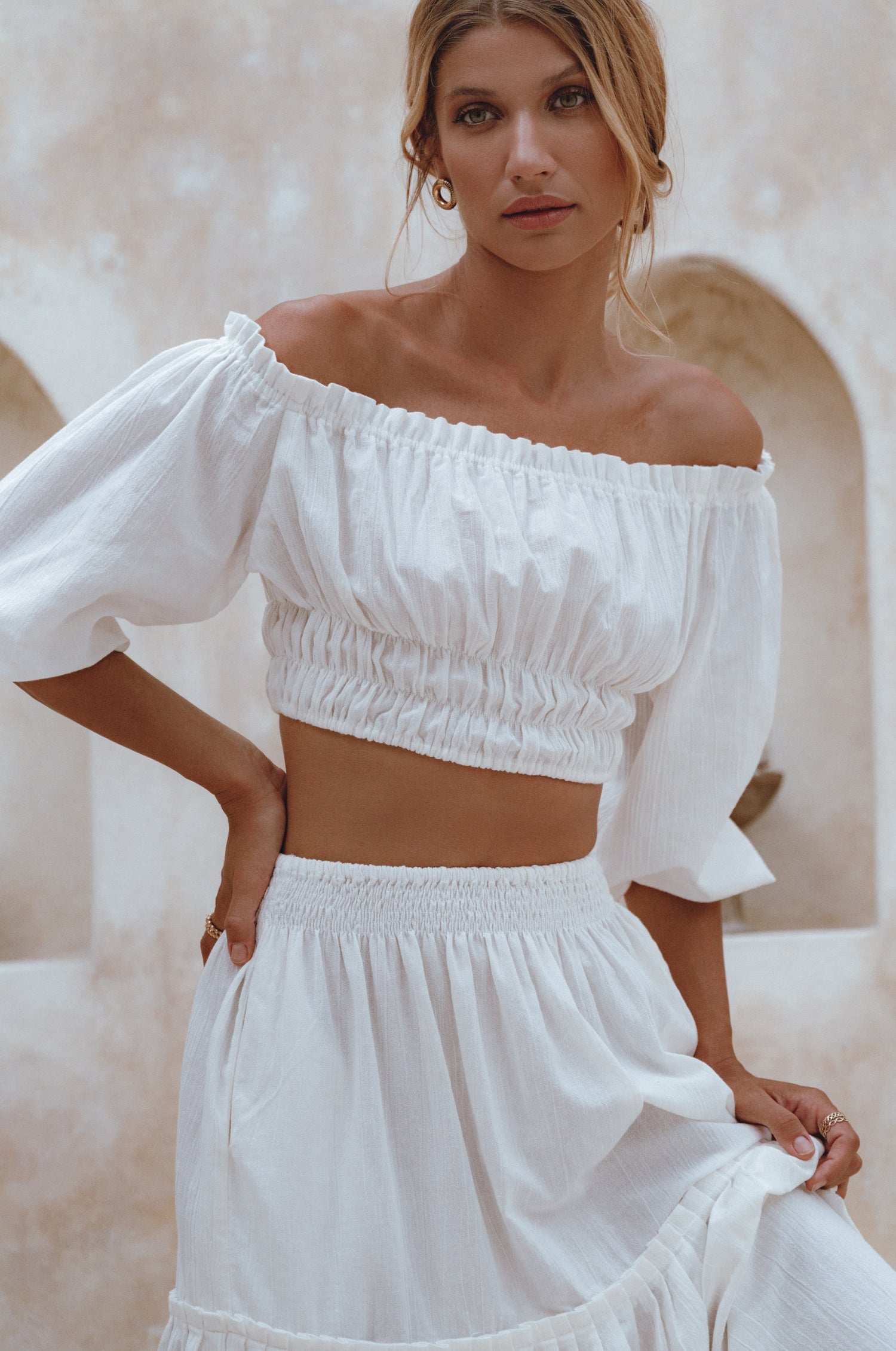 Woman wearing a white linen cropped top with elasticated detailing at the waist and neckline worn off-the-shoulder against a white background, front view