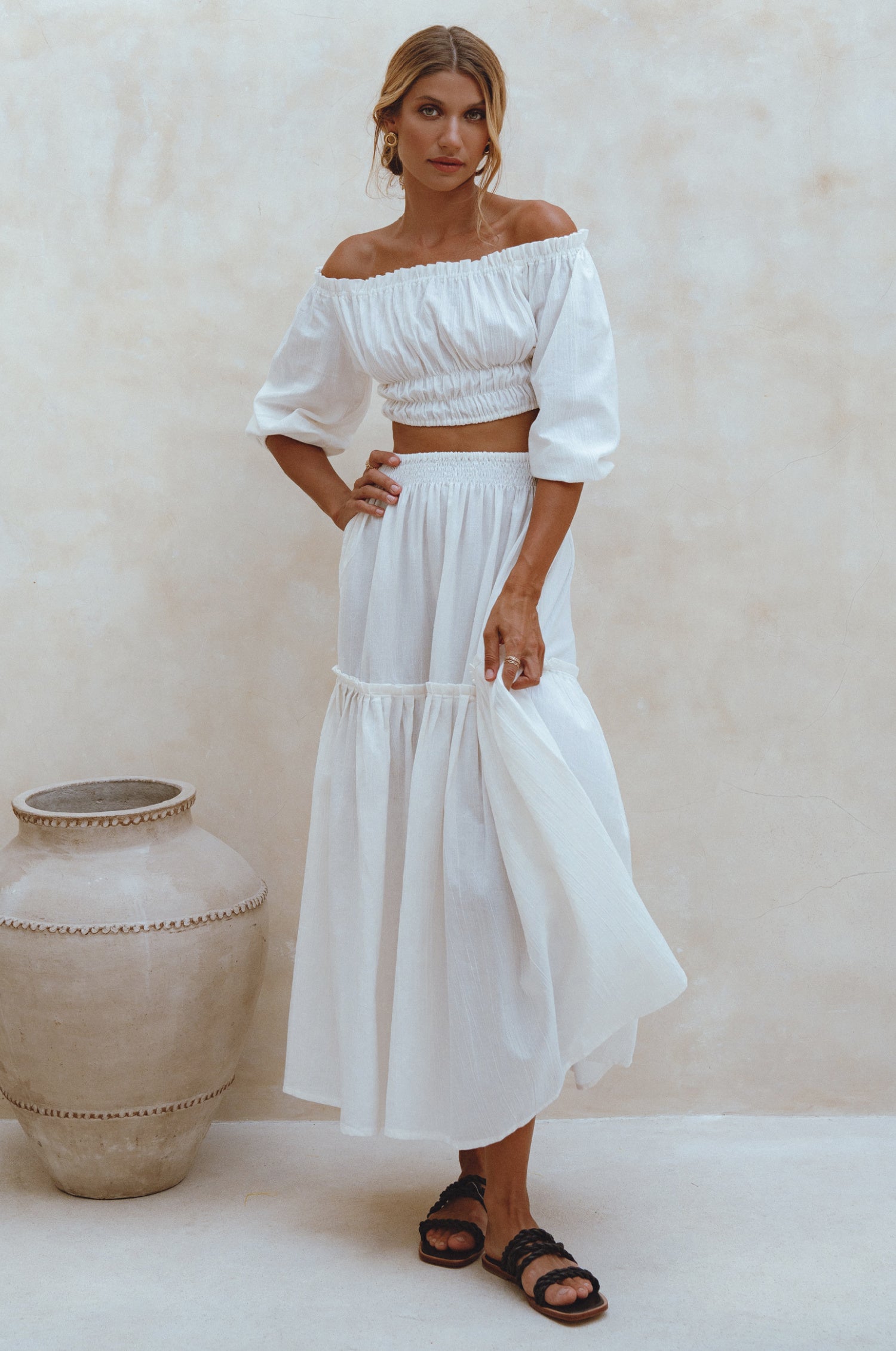 Woman wearing white crinkle linen high-waist two-tier maxi skirt and matching cropped off-shoulder blouse against neutral background, front view. 