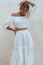 Woman wearing white crinkle linen high-waist two-tier maxi skirt and matching cropped off-shoulder blouse against neutral background, front view. 