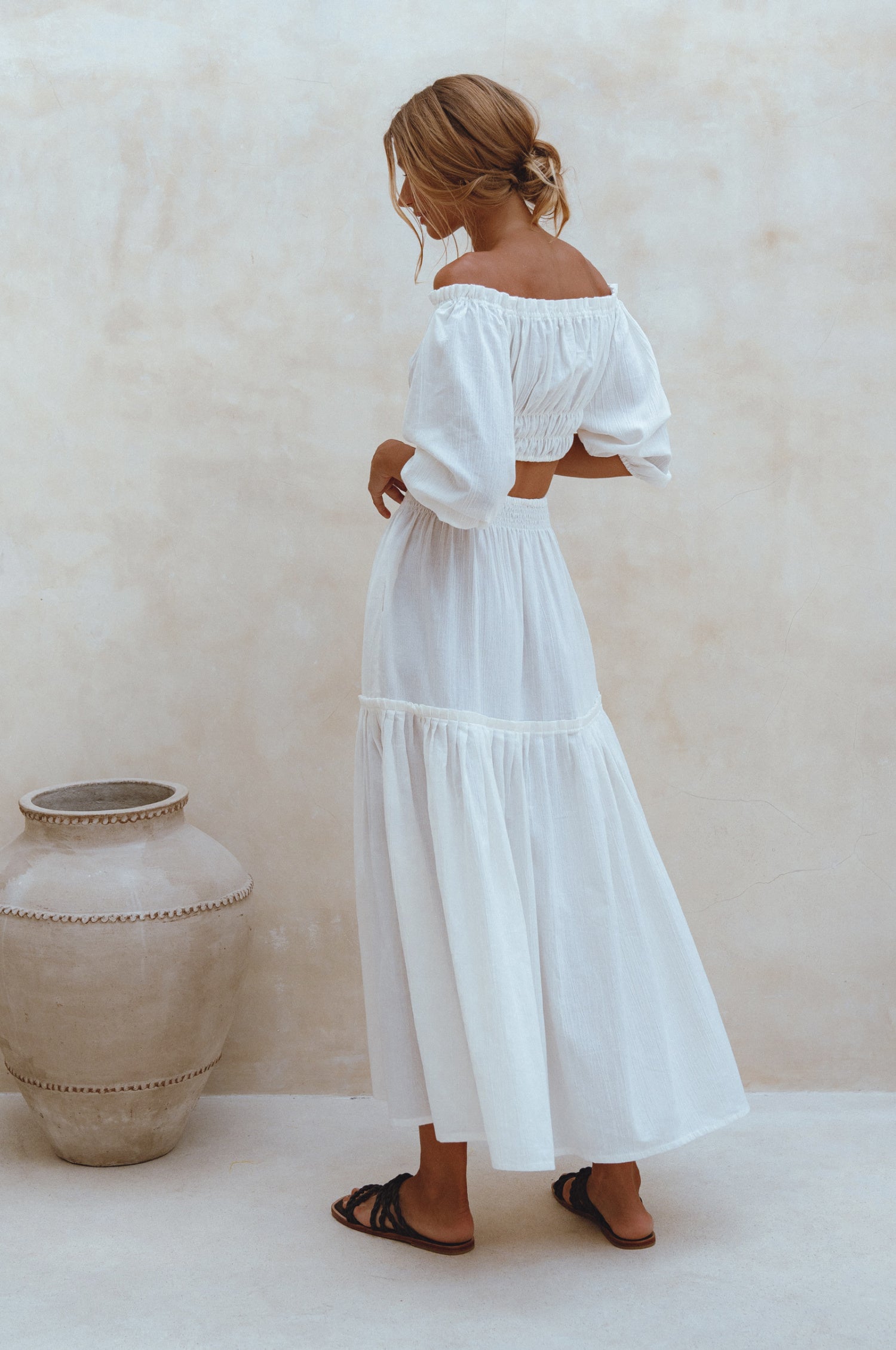 Woman wearing white crinkle linen high-waist two-tier maxi skirt and matching cropped off-shoulder blouse against neutral background, back view. 