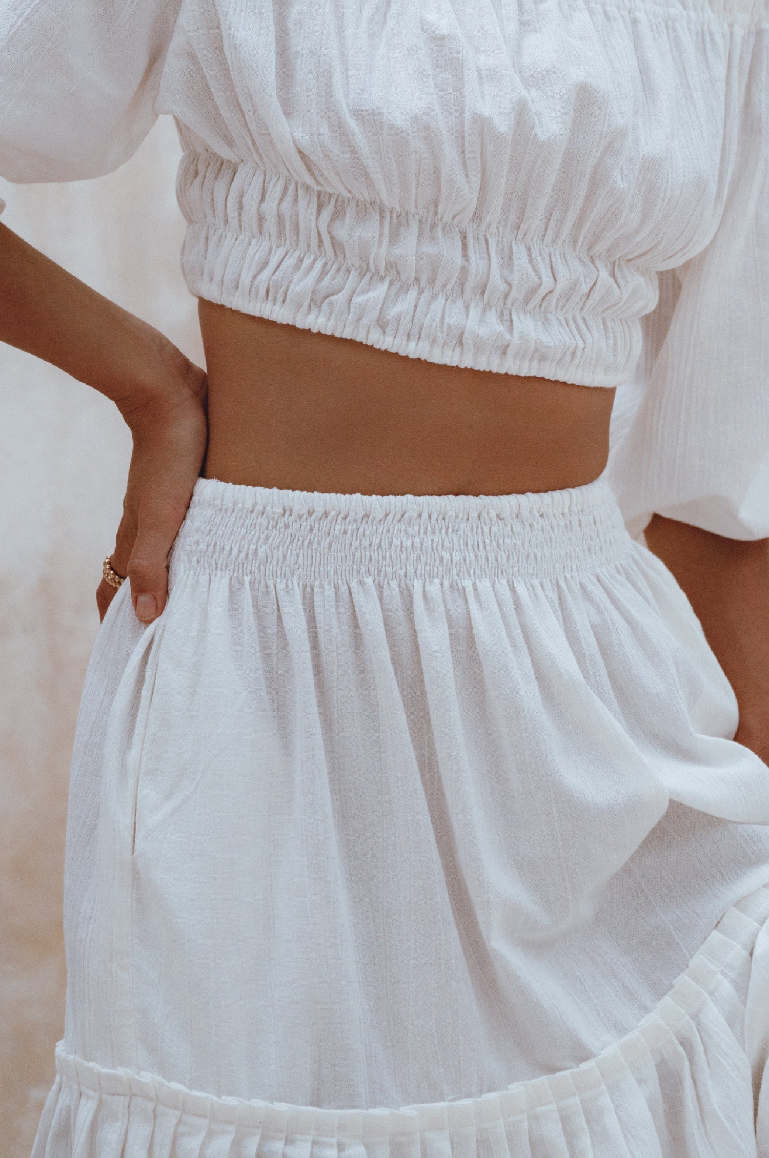 Woman wearing white crinkle linen high-waist two-tier maxi skirt and matching cropped off-shoulder blouse against neutral background, front view close up. 