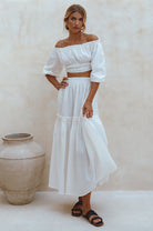 Woman wearing white crinkle linen high-waist two-tier maxi skirt and matching cropped off-shoulder blouse against neutral background, front view. 