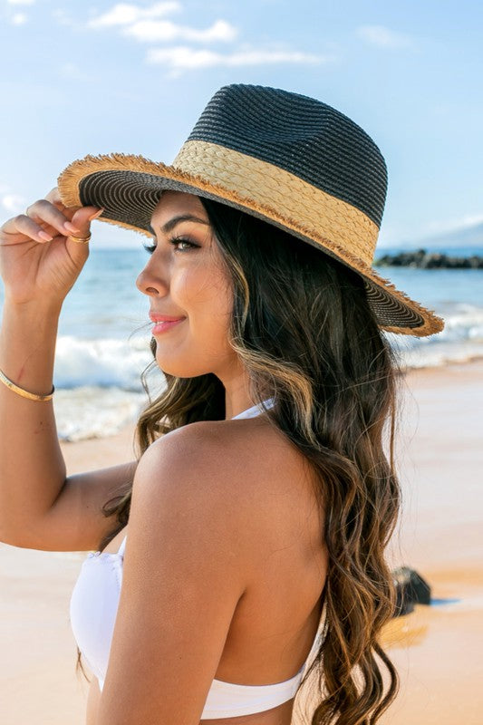 Side view of woman wearing a black straw sun hat with natural rattan band and holding the frayed fringe brim at the beach