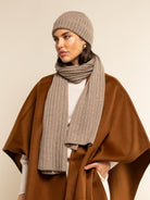 Woman wearing a brown coat, beige beanie, and scarf against a beige background