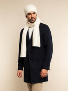 Man wearing a navy coat, ivory scarf, and beanie against a beige background