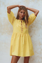 Woman wearing a yellow linen mini dress with puff sleeves standing against a textured white wall, front view