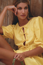 Woman wearing a yellow linen mini dress with puff sleeves and ties in the front bodice, leaning against a wood wall, front view closeup